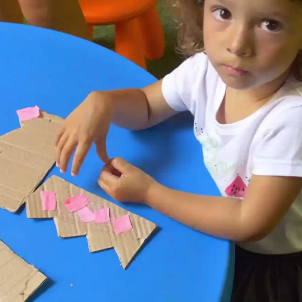 Ein Kind bastelt mit Karton und rosa Papier an einem blauen Tisch.