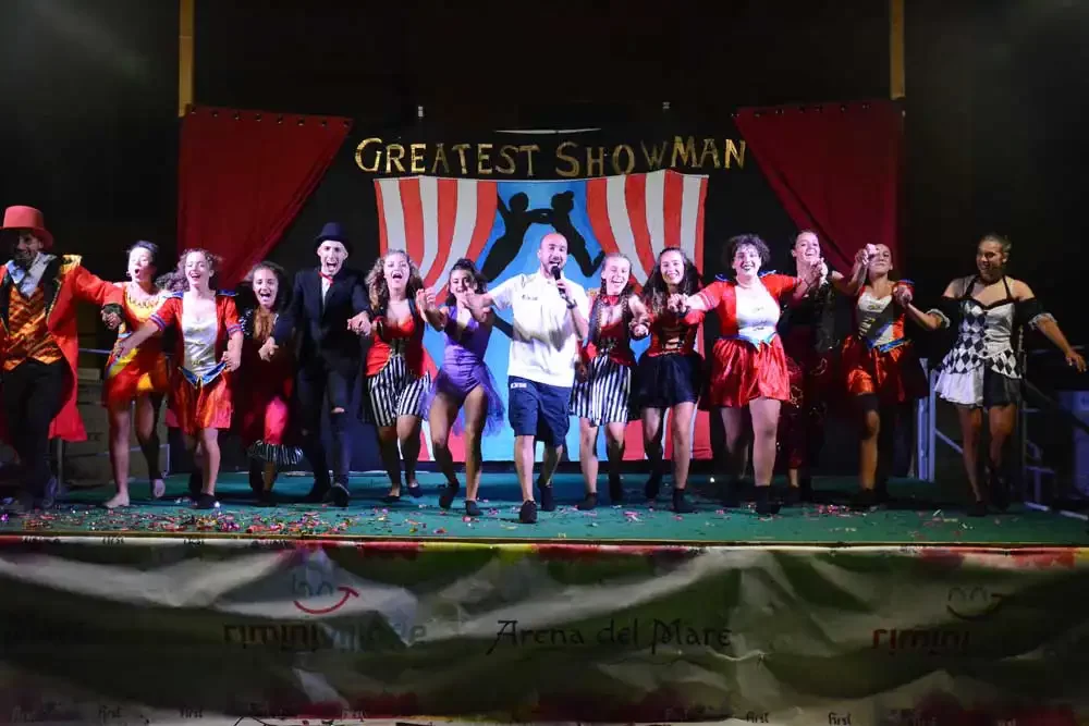 Gruppe von Menschen in bunten Kostümen bei einer Theateraufführung mit dem Titel "Greatest Showman" auf der Bühne.
