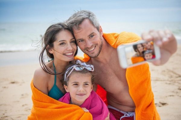 Un couple souriant avec un enfant se prend en selfie sur la plage, enveloppé dans des serviettes colorées.